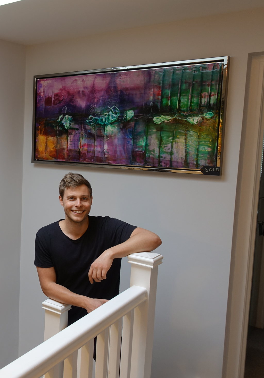 Max Argyle standing beside a hang of his framed mixed-media paintings